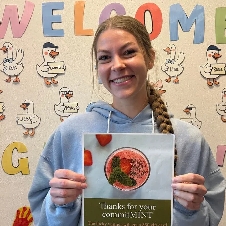 Miss Lexi smiling and holding a thank-you card in front of a classroom welcome sign
